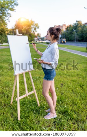 beautiful girl city summer Full length, woman artist, draws picture road, beginning of drawing, first brush stroke, white canvas painting, easel stand. Green grass lawn background, cars on highway