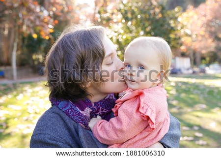 Happy mother kissing baby girl in cheek with closed eyes. Portrait of attractive Caucasian woman walking in autumn park with adorable girl sitting on mother hands. Family and maternity concept