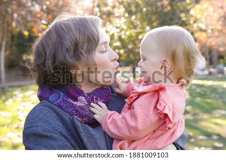 Side view of mum holding little daughter and kissing her with closed eyes. Portrait of beautiful blonde woman standing in autumn park. Cute girl sitting on mother hands. Family and motherhood concept