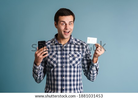 Photo of happy young man isolated over blue wall background holding mobile phone and credit card, celebrating