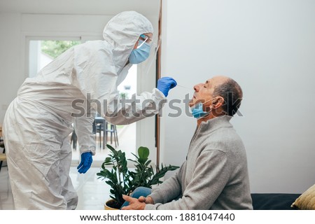 Health Professional in PPE suit and face shield introducing a nasal swab to a senior adult patient at his house. Rapid Antigen Test kit to analyze nasal culture sampling while coronavirus Pandemic.