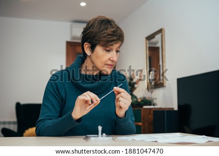 Senior woman sitting at home, studying the instructions for a self test for COVID-19 with Antigen kit on hand. Holds a nasal swab for possible infection of Coronavirus. Health services online.
