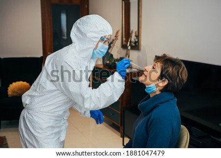 Health Professional in PPE, face mask and shield, introducing a nasal swab to a senior female patient on a home visit. Rapid Antigen Test to analyze nasal sampling for Covid-19, Coronavirus Pandemic.