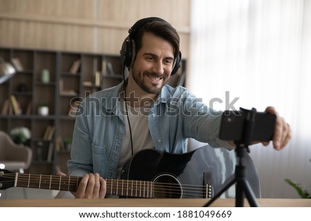 Smiling millennial male artist blogger playing on guitar shooting music video or tutorial on modern smartphone at home studio. Happy young man singer use music instrument, compose song on cellphone. Royalty-Free Stock Photo #1881049669