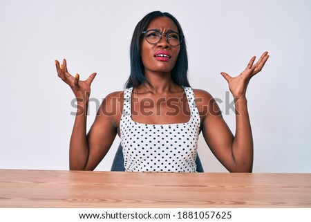 Young african american woman wearing casual clothes and glasses sitting on the table crazy and mad shouting and yelling with aggressive expression and arms raised. frustration concept. 