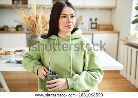 Indoor shot of stylish dark haired young Caucasian woman in oversized hoodie sitting in kitchen with mug, drinking coffee or hot chocolate, having pensive facial expression, thinking about plans