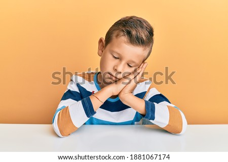 Adorable caucasian kid wearing casual clothes sitting on the table sleeping tired dreaming and posing with hands together while smiling with closed eyes. 