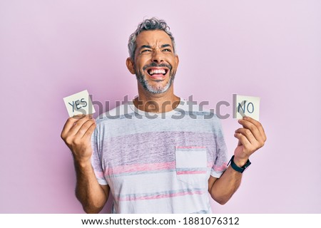 Middle age grey-haired man holding yes and no reminder smiling and laughing hard out loud because funny crazy joke. 