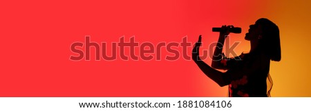 Flyer. Silhouette of young female singer isolated on orange gradient studio background in neon light. Beautiful shadow in action, performing. Concept of human emotions, expression, ad, music, art. Royalty-Free Stock Photo #1881084106
