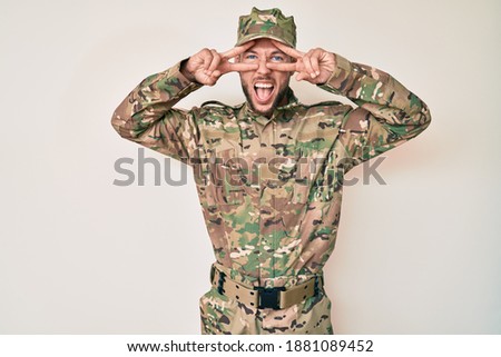 Young caucasian man wearing camouflage army uniform doing peace symbol with fingers over face, smiling cheerful showing victory 