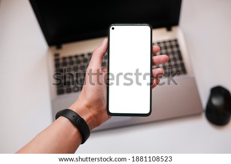 Close-up of a smartphone with a white screen in hand. Laptop, mouse, selective focus, internet business and finance concept with phone with copy space, top view.