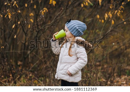 A little girl warmly dressed preschool age stands in nature and drinks delicious and hot tea from a plastic green cup, basking in the cold. Autumn portrait in October, photography.