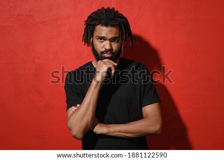 Pensive puzzled serious young african american man with dreadlocks 20s wearing black casual t-shirt posing put hand prop up on chin looking camera isolated on red color wall background studio portrait