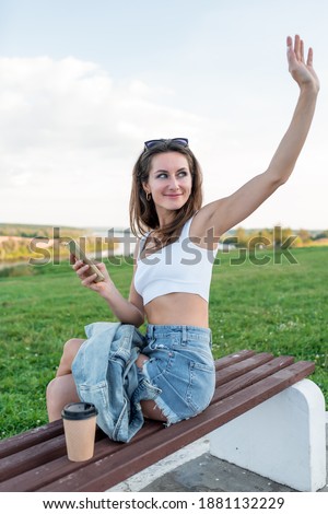 woman in city in summer, sits on a bench, waving to her girlfriends and friends. Happy smiles, sends greetings. Holds a smartphone in his hand. Background green grass river and clouds