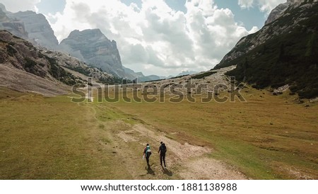 A couple hiking along a pathway leading through a valley in Italian Dolomites. High, sharp mountains around. Stony and raw landscape, green meadow. Remote and desolate place. Freedom of exploration