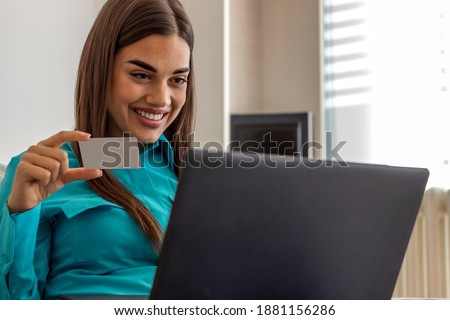 Smiling young woman holding a credit card and typing on a laptop. Photo of a joyful young girl doing online shopping while sitting on couch at home during the day. Woman shopping online in living room