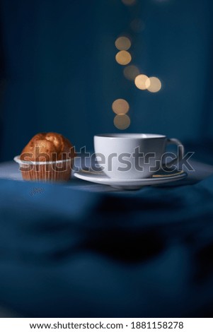 Tea in white cup with fresh cupcake on the blue background with blurred lights in the cafe