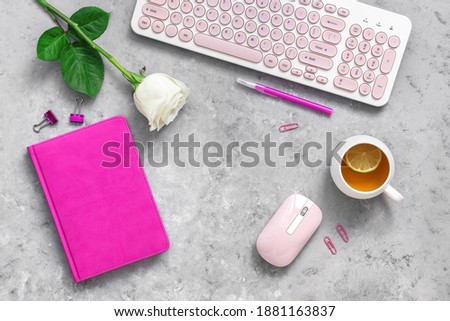 Feminine freelance workspace concept. Workplace of a blogger or office worker. Keyboard, blank notepad, mouse, cup of tea and rose flower. Top view, flat lay. Gray concrete grunge background