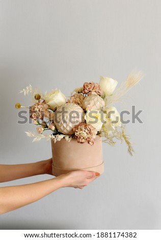 Flowers for valentines day. Floral composition. Fresh present of bouquet and holiday decor: balls, dried flowers in hands of a florist woman. Cylindrical box on grey showcase shop background.