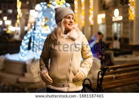 Outdoor photo of young beautiful happy smiling girl holding sparklers, posing in street. Festive Christmas fair on background. Model wearing stylish winter coat, knitted beanie hat, scarf.