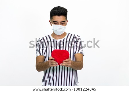 Young man holding a heart shaped gift box in his hand. He wears a medical mask on his face. Wearing a striped T-shirt. isolated image, white background