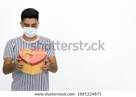 Young man holding a heart shaped gift box in his hand. He wears a medical mask on his face. Wearing a striped T-shirt. isolated image, white background