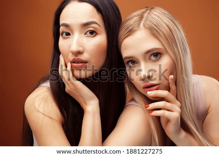two pretty diverse girls happy posing together: blond and brunette on brown background, asian and caucasian, lifestyle people concept