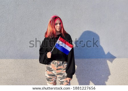 Student girl teenager with the flag of the Netherlands in hand, gray outdoor wall background. Europe, Netherlands, education and youth, patriotism people concept