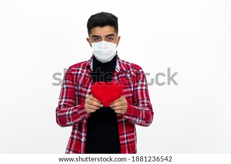 Young male wearing a medical mask. The man is holding a heart-shaped gift box in his hand. Wearing a red, striped shirt. White background, isolated image.