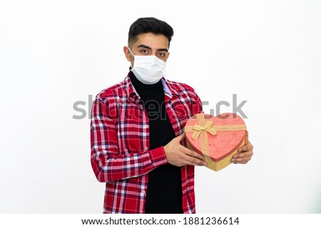 Young male wearing a medical mask. The man is holding a heart-shaped gift box in his hand. Wearing a red, striped shirt. White background, isolated image.