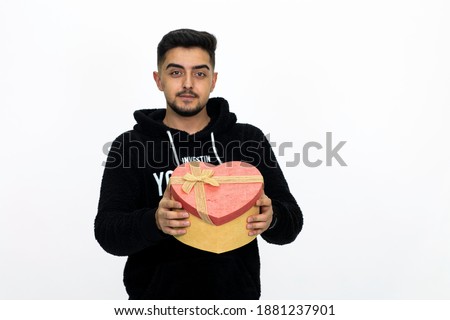 Young male holding a heart-shaped gift box in his hand. He is dressed in a black hooded sweater. White background, isolated image.