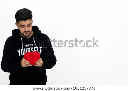 Young male holding a heart-shaped gift box in his hand. He is dressed in a black hooded sweater. White background, isolated image.