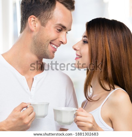 Happy smiling amazed couple with cups of coffee or some drinks at home. Portrait of standing close and looking at each other models in love family concept. Man and woman posing together. 