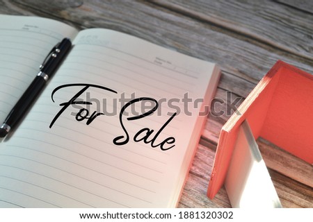 Selective focus of pen, toy wooden house and notebook written with text FOR SALE over wooden background.