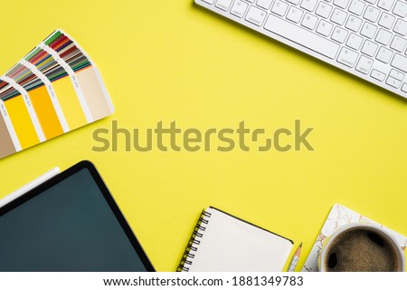 Graphic designer desk top view with RAL colors palette, tablet, notebook, cup of coffee, computer keyboard on yellow background. Flat lay, view from above. Professional creative workspace.