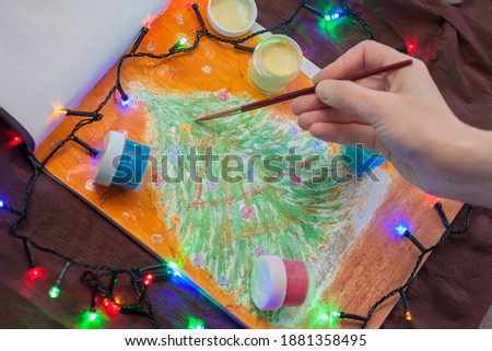 Female's hands holding the brush and Painting beautiful Christmas tree. There are blue, yellow, red, green paintings and the colorful garland near the picture. New year (Christmas) background