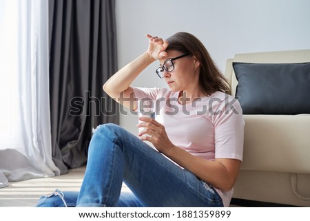 Serious sad middle aged mature woman sitting at home holding glass of water looking out the window. Health problems, mental difficulties, depression, menopause, and personal troubles Royalty-Free Stock Photo #1881359899