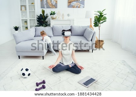 Mother meditating in lotus yoga position uses vr goggles while her daughter watches cartoons on background. Top view