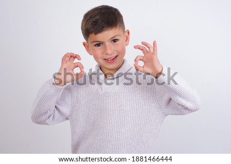 Cute Caucasian kid boy wearing knitted sweater against white wall showing both hands with fingers in OK sign. Approval or recommending concept