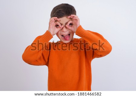 Cute Caucasian kid boy wearing knitted sweater against white wall doing ok gesture like binoculars sticking tongue out, eyes looking through fingers. Crazy expression.