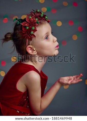 Christmas kiss. Kid girl wearing red sequin dress and Christmas wreath. Blond hair and blue eyes posing and fool around in studio on grey background with illumination and boke lights.