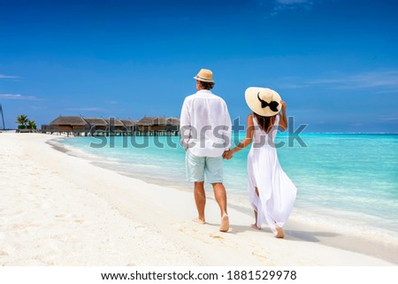 Happy couple in white clothing and with hats walks down a tropical beach with turquoise sea in the Maldives islands Royalty-Free Stock Photo #1881529978