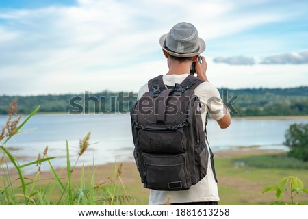 Young Asian man traveler wearing knitted hat with backpack taking a photo and looking at amazing lake. Travel holiday relaxation concept