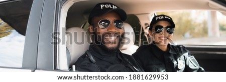 happy multicultural police officers looking at camera in patrol car on blurred background, banner