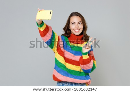 Smiling cheerful young brunette woman 20s wearing casual colorful knitted sweater standing doing selfie shot on mobile phone showing victory sign isolated on grey colour background, studio portrait