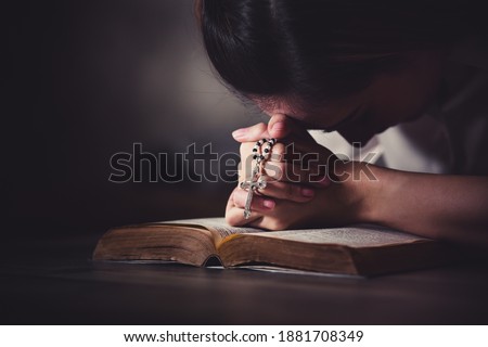 Christian Asian woman praying and holding rosary cross on bible. Close up casual female hands praying with her hands over open bible with beautiful light in darkened room. Royalty-Free Stock Photo #1881708349