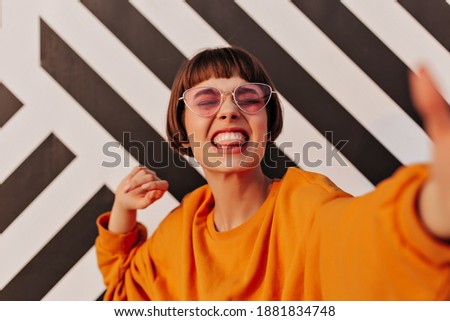 Emotional woman with short hairstyle in pink glasses showing tongue on striped backdrop. Modern girl in orange sweatshirt makes funny face outside..