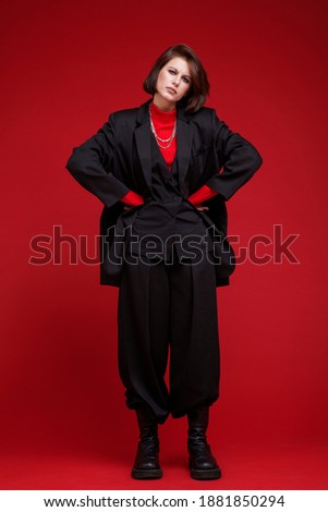 Fashion photo of a beautiful elegant young woman in a pretty oversize black suit, pants, top posing over red background. Bob haircut. Studio Shot. Rebellious free street style. Rebel and classic
