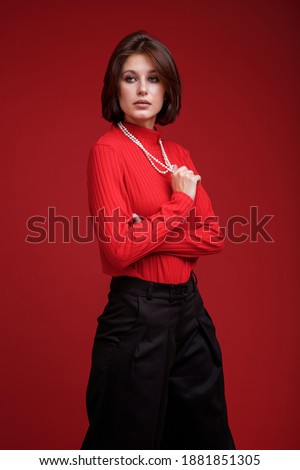 Fashion photo of a beautiful elegant young woman in a pretty oversize black pants, red top, pearl necklace posing over red background. Bob haircut. Studio Shot. Black and red colors