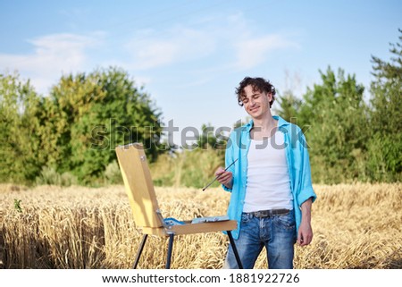 Young male artist wearing light blue shirt, drawing on canvas on sketchbook easel on wheat field. Painting workshop in rural countryside. Artistic education concept. Outdoors leisure activities.